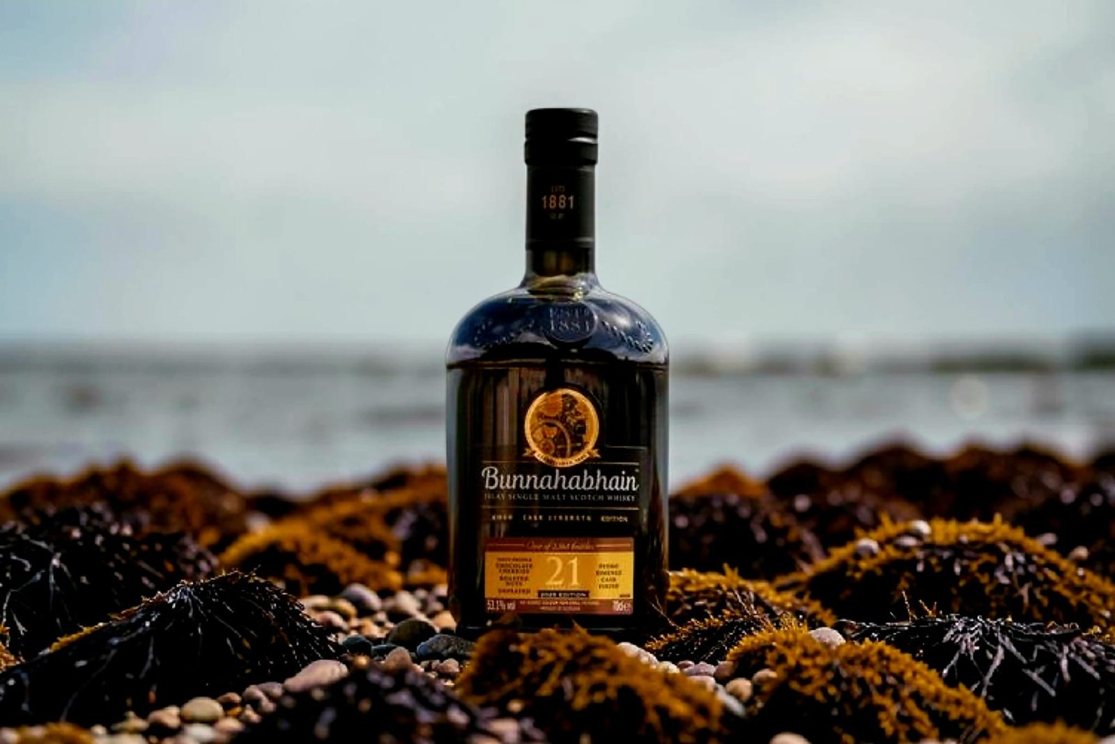 Bunnahabhain 21 Cask Strength 2025 whisky bottle on Islay shoreline with sea and rocks