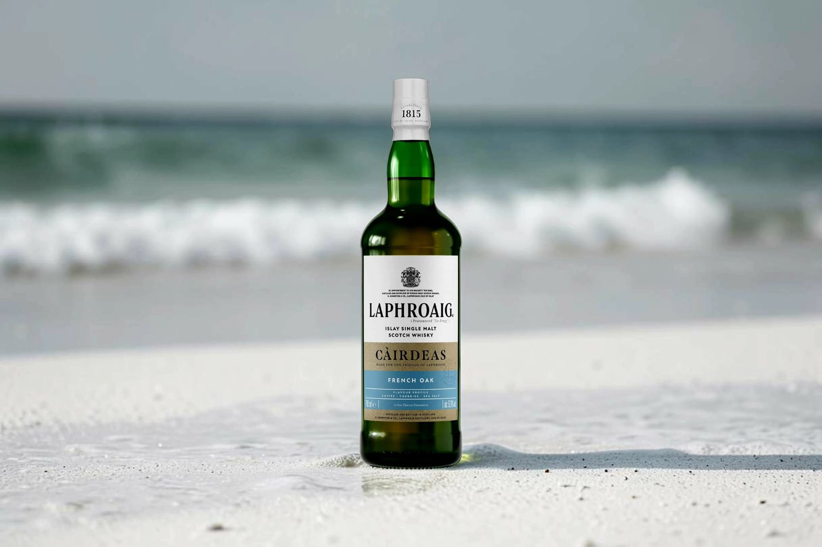 Laphroaig Càirdeas 2026 French Oak bottle standing on a sandy beach with ocean waves breaking in the background.